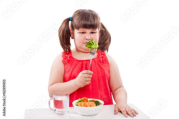 Fototapeta Overweight child sitting at the table and looking unhappily at a fork with broccoli. Concept of unhealthy eating habits, food rejection, childhood obesity, and picky eating. White background.