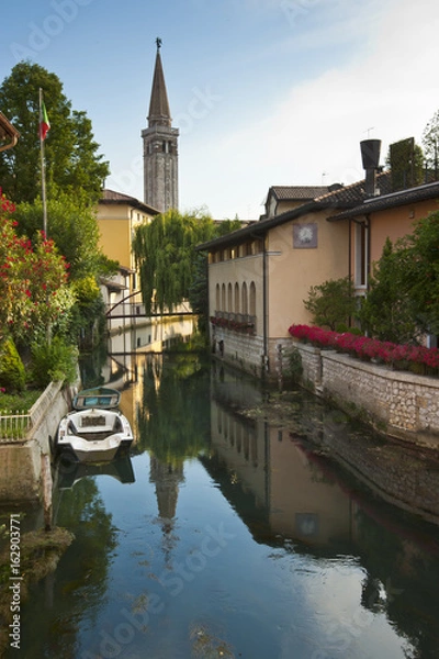 Fototapeta The tower bell of the cathedral of St. Nicolò is reflected on Livenza river in Sacile. Italy