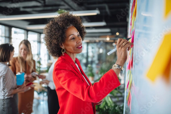 Fototapeta Mature Black business woman collaborating in an open-space office, brainstorming on sticky notes placed across a whiteboard, wearing a fashionable red blazer, actively engaged with colleagues, modern
