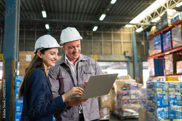 Fototapeta Warehouse Manager  and supervisor in inventory data on a laptop amid stacked pallets.