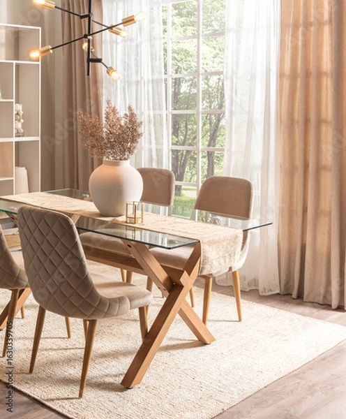 Fototapeta Spacious dining room in Nordic style showcasing a sleek glass tabletop, fabric quilted chairs, an intricate chandelier, and ambient sunlight filtering through sheer curtains, on a woven area rug.