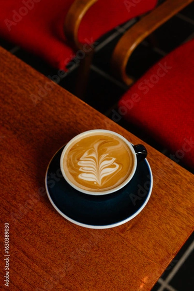 Fototapeta Cappuccino with plant based milk in a large mug on a wooden table. A cup of coffee against the background of cozy red armchairs. Specialty coffee concept