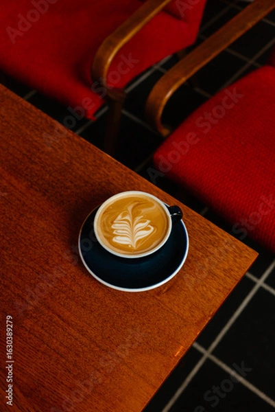 Fototapeta Cappuccino with plant based milk in a large mug on a wooden table. A cup of coffee against the background of cozy red armchairs. Specialty coffee concept