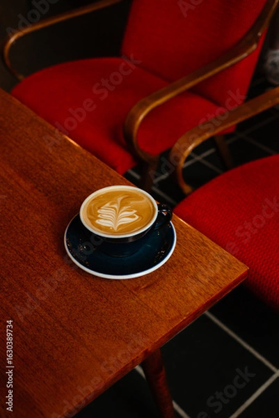 Fototapeta Cappuccino with plant based milk in a large mug on a wooden table. A cup of coffee against the background of cozy red armchairs. Specialty coffee concept