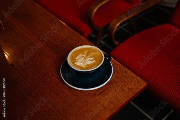 Fototapeta Flat white in a large cup on a wooden table next to red armchairs. Coffee break in a special coffee shop. Aromatic coffee with milk for breakfast