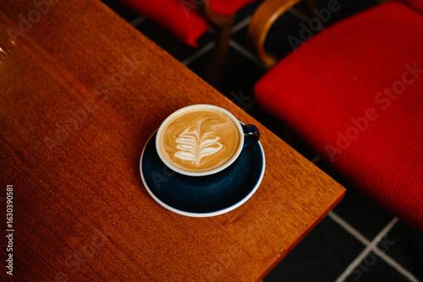 Fototapeta Flat white in a large cup on a wooden table next to red armchairs. Coffee break in a special coffee shop. Aromatic coffee with milk for breakfast