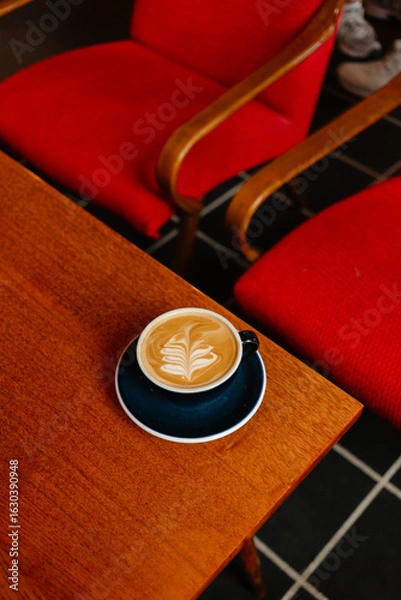 Fototapeta Flat white in a large cup on a wooden table next to red armchairs. Coffee break in a special coffee shop. Aromatic coffee with milk for breakfast