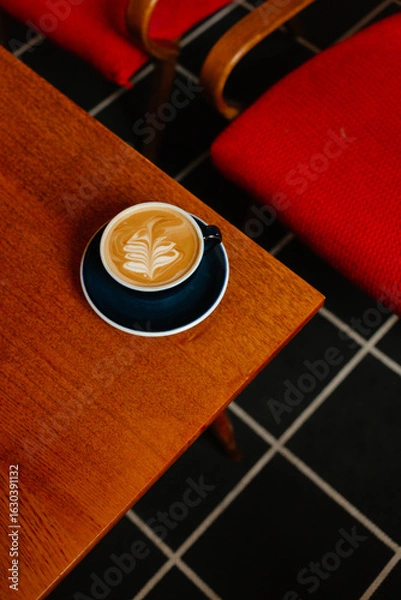 Fototapeta Flat white in a large cup on a wooden table next to red armchairs. Coffee break in a special coffee shop. Aromatic coffee with milk for breakfast