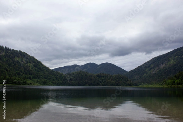 Obraz Serene mountain lake under a dramatic overcast sky, surrounded by lush green forested hills. The calm water reflects the surrounding nature, creating a peaceful and moody atmosphere 