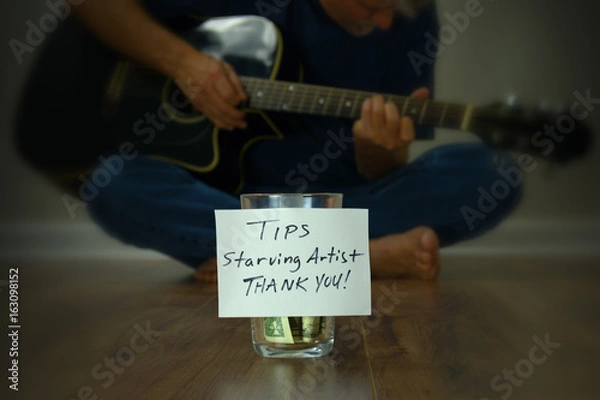 Fototapeta Starving artist guitarist street performer sitting on the sidewalk playing his guitar with a tip cup jar in front of him in crisp focus.