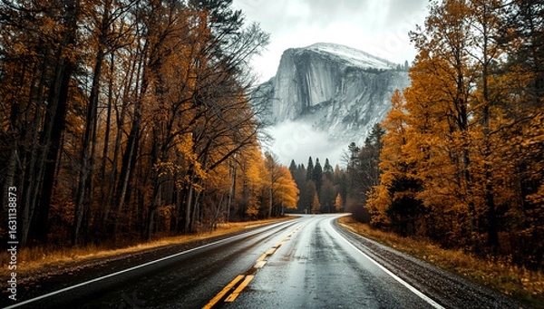 Fototapeta A winding road leads through a forest of autumn trees with orange and brown leaves, towards a misty, rocky mountain in the background.