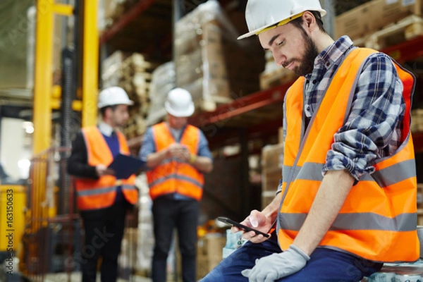 Fototapeta Man in uniform and helmet messaging at break in warehouse