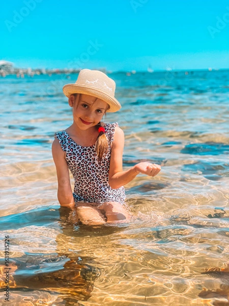 Obraz A young girl in a hat sits in shallow water on a sandy beach, surrounded by clear turquoise waves. Peaceful summer scene with bright sunlight, relaxing atmosphere, and tropical seaside vibes.
