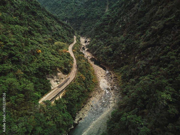 Obraz Aerial view of a winding road through a lush, mountainous valley with a river running through it.