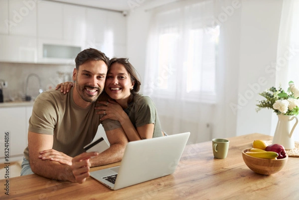 Fototapeta Happy couple using credit card for online shopping over laptop at home and looking at camera.