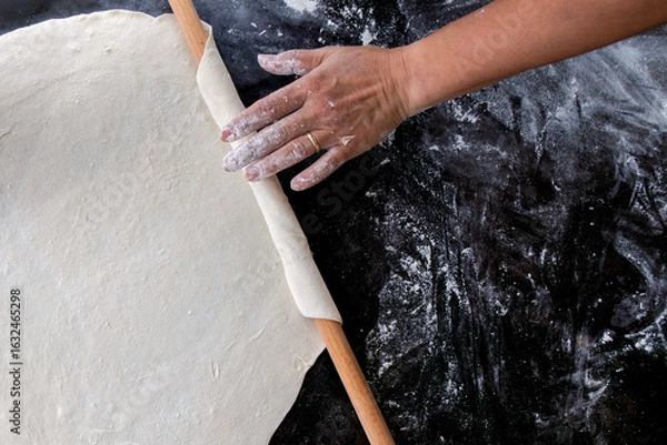 Fototapeta A top-down view of a hand rolling out dough with a wooden rolling pin on a dark, floured surface. 