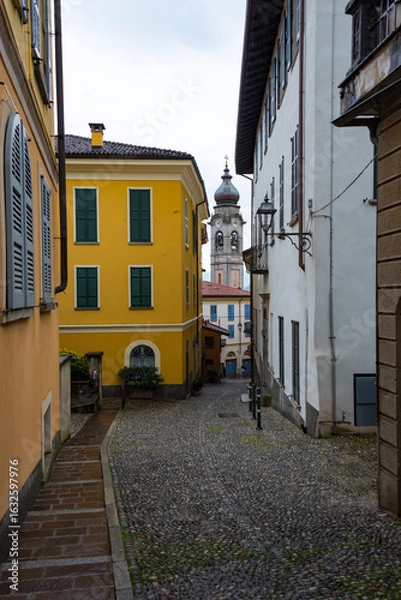 Fototapeta A narrow road paved with round stones, old buildings with shutters and church in a gap between them in Menaggio, Lombardia, Italy