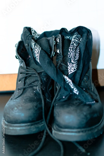 Fototapeta Pair of black steel toe work boots on a table in front of a dry erase board calendar. The boots also have a black and white bandanna wrapped around the top portion.