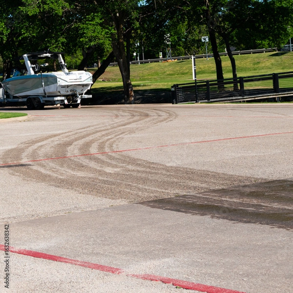 Fototapeta A white an grey boat in the distance following a trail of wet tire marks from recently pulling the boat out of the lake on a sunny evening. 