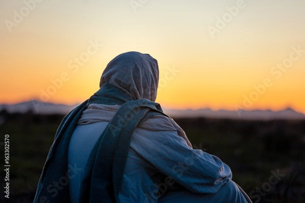 Fototapeta Jezus Chrystus siedzi na zboczu wzgórza i obserwuje słońce zachodzące na horyzoncie.