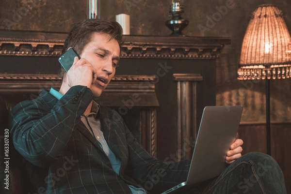 Obraz Man with computer sitting by the fireplace and talking on the phone