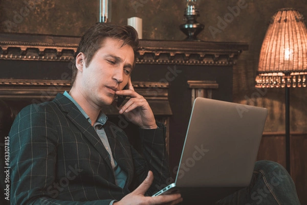 Obraz Man with computer sitting by the fireplace and talking on the phone
