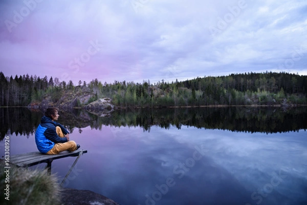 Fototapeta Man sitting on a pier at mountain lake, looking at beautiful at sunset. Scandinavia travel destination.