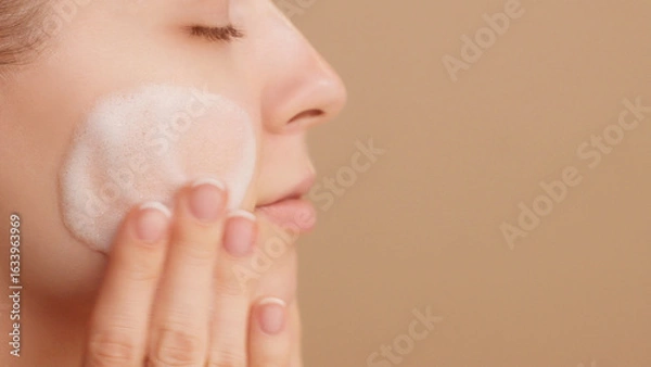 Fototapeta Close-up of a beautiful female face. Woman applying cleansing foam gently washing face ads. Bubbles. Beauty daily routine. Commercial shoot. Skincare treatment. Selfcare. Copy space. Beige Background