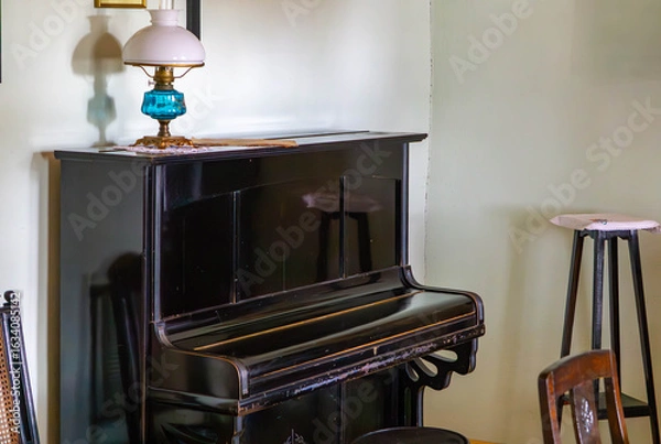 Fototapeta Vintage upright piano against a wall in a classic interior, with an ornate kerosene lamp placed on top. A chair and a small stool nearby.
