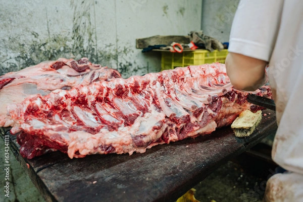 Obraz A butcher cleans a large rack of raw beef ribs on a wooden cutting board in a dimly lit, industrial setting.