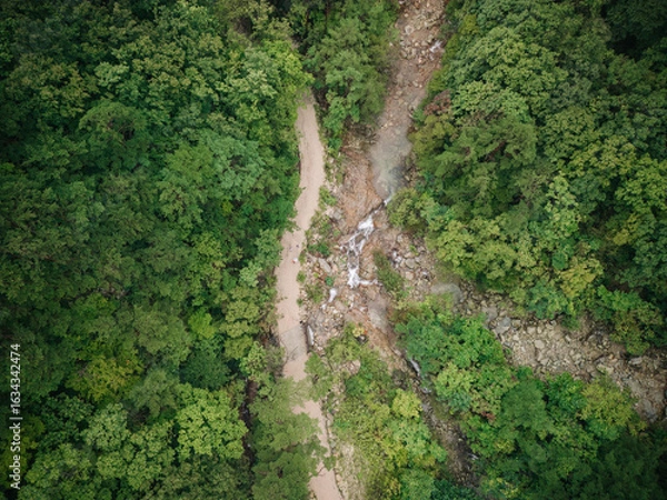 Obraz Aerial view of a dirt path winding through a lush, forested ravine with a small waterfall.