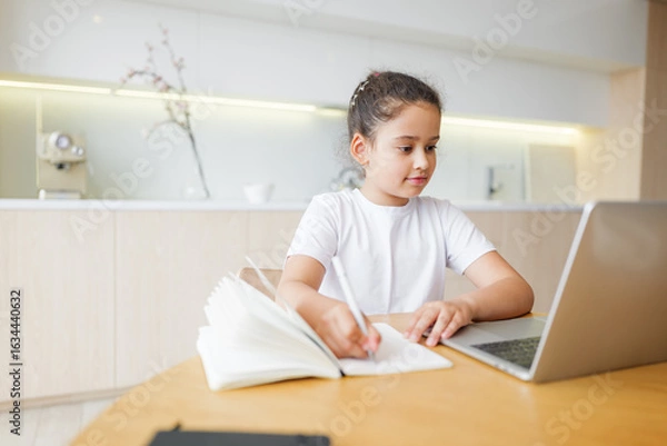 Fototapeta Young girl engaged in online learning, balancing note-taking and digital education in a modern kitchen setting, fostering creativity and growth