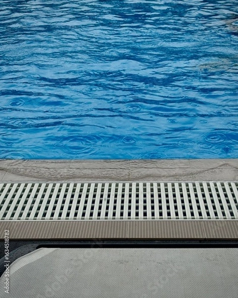 Fototapeta Blue swimming pool water with rain droplets and ripples near lounge chair on a cloudy day. 