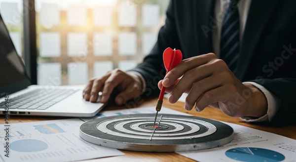 Fototapeta Businessman Achieving Goals:  Close-up Shot of Hands Placing Dart in Bullseye on Dartboard, Laptop and Financial Charts Present.