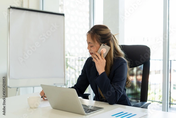 Fototapeta Businesswoman sitting at desk on couch in workplace or at home working on laptop and analyzing data on charts and graphs and writing on papers to make business plan and strategies for company.