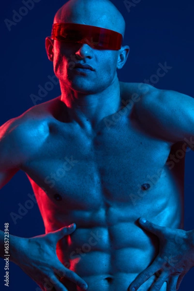 Fototapeta Athletic man posing confidently with sunglasses, showcasing his toned physique in vibrant blue and red lighting. A striking black and white aesthetic enhances the impact.