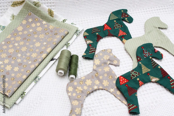 Fototapeta Christmas tree toys made by hand. On a flat surface beautiful cotton fabric with a New Year's ornament, handmade horses and threads. White background.