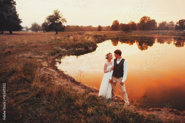 Fototapeta The bride and groom posing against the background of the lake in the red sunset. Overall plan