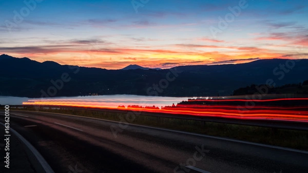 Fototapeta Austrian countryside road car is passing during the sunset only light rays are visible lacke is on the background