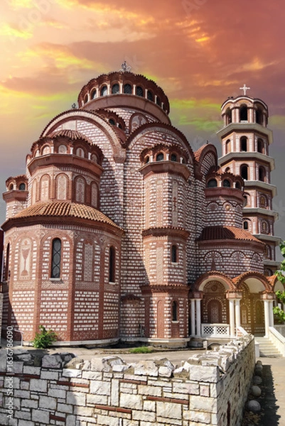 Fototapeta The magnificent Holy Church of Panagia Korifini in Nea Moudania, Halkidiki, showcasing stunning Byzantine architecture with intricate red brickwork against a dramatic and colorful sunset sky in Greece