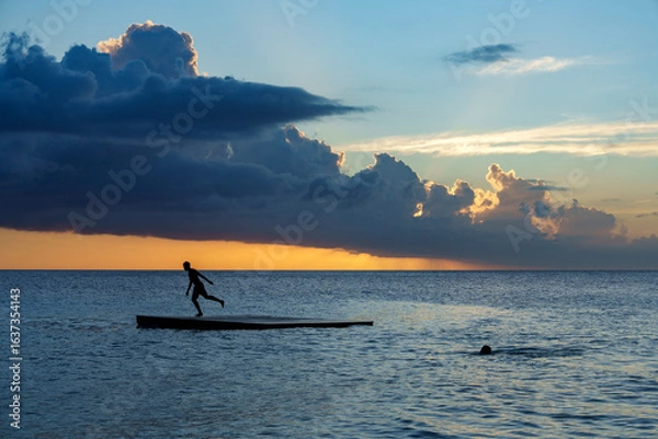 Obraz Crianças brincando em plataforma flutuante no mar do Caribe ao pôr do sol em Curaçao, capturando momentos de diversão, liberdade e beleza tropical.