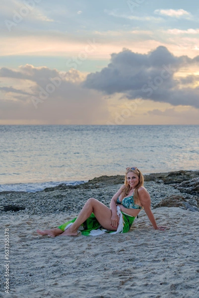 Fototapeta Beautiful woman in colorful sarong relaxing on a tropical Caribbean beach in Curaçao at sunset, with golden light, turquoise water, and serene holiday vibes