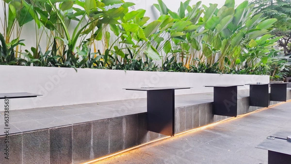 Obraz Cozy modern outdoor cefe with black metal table, lush tropical plants in white pot, and urban building backdrop. Perfect for relaxaxing and enjoying coffee. peacefull ambient.