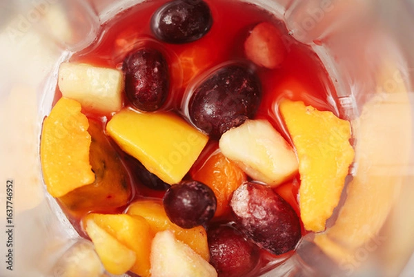 Fototapeta Fresh fruit pieces in blender. Top view of mango, pineapple, cherries and grapes with red juice. Ingredients prepared for smoothie or juice. Healthy natural drink raw ripe tropical and berry fruits.