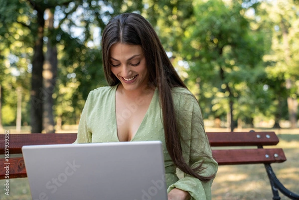 Fototapeta Beautiful smiling young woman working on laptop in park sitting on bench enjoying daylight and fresh air