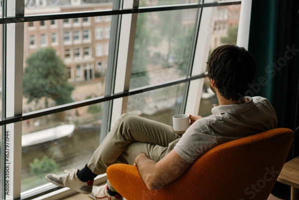 Fototapeta man drinking coffee in a chair with attic window and view on Amsterdam	