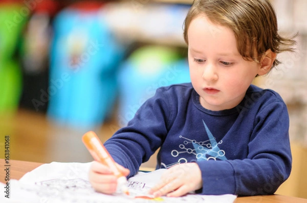 Fototapeta Young boy sitting down at desk indoors coloring with markers
