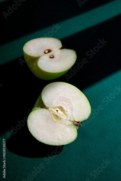 Obraz Minimalist composition of a fresh green apple cut in half, placed on a deep green surface with dramatic shadows. 