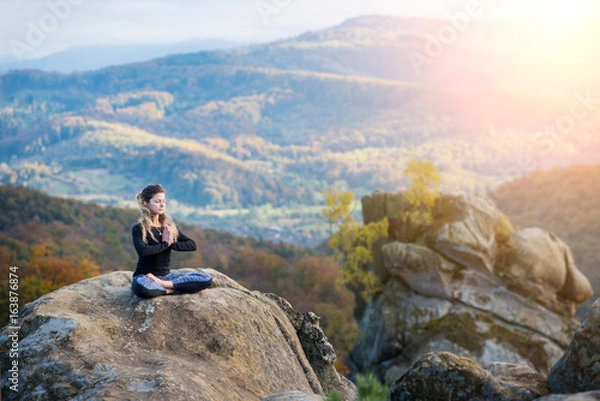 Fototapeta Pretty young woman is practicing yoga and doing asana Siddhasana on the top of the mountain in the evening. Autumn forests, rocks and hills on the background