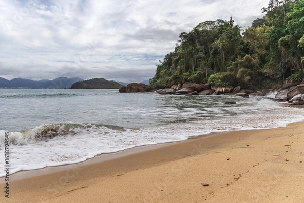 Fototapeta Ubatuba beach in São Paulo, with trees at the background, sand and waves.
Incredible paradisiac beach with cloudy sky.
